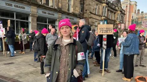 BBC Jo Grady berdiri di depan sekelompok piket. Dia mengenakan topi merah muda yang berbunyi: "UCU". Di belakangnya, sebuah plakat berbunyi: "Jangan silang piket".