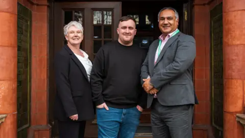 Warrington and Halton Teaching Hospitals Charity Three people stand outside a door with ornate red brick columns and green tiled panels. Luke Littler and two hospital officials are wearing suits. Littler wears a casual black sweatshirt with jeans.