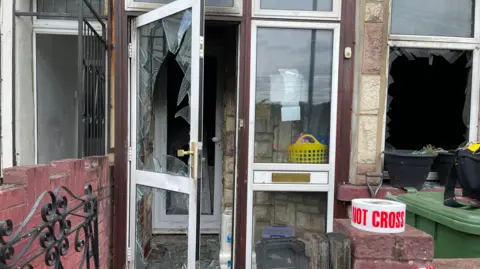 Front door of the house, with the glass sections smashed in. There's a black iron gate into the front garden area, and some red and white 'do not cross' tape hanging on the low brick wall