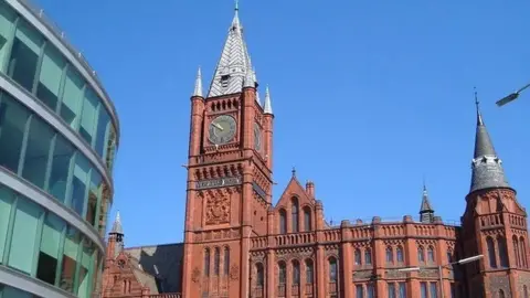 A red brick building with a clock tower behind a modern glass curved building