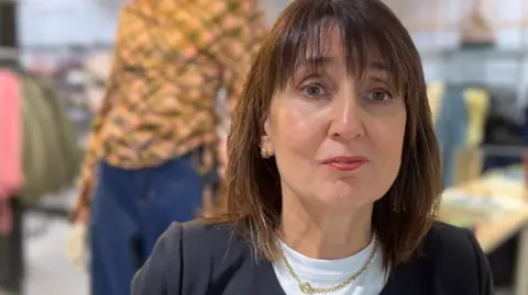 Tracey Odell is wearing a black cardigan over a white T-shirt. She has short brown hair with a fringe. Clothes are hanging on rails behind her and there is a mannequin wearing a colourful orange, green and black top and blue jeans.