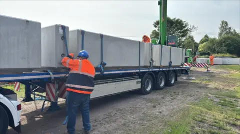 Gary Essex A man in an orange hi-vis jacket and blue helmet is pulling a blue strap. The strap is tied around a large block of concrete and is secured to a long trailer.