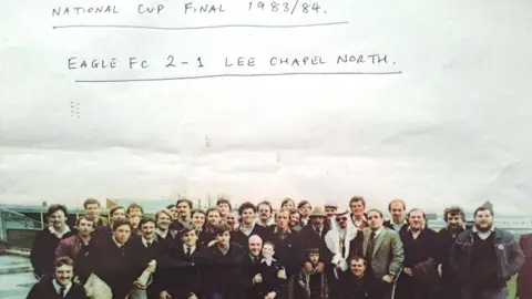 John Burns A group of men have gathered for this group photographs. Written on the top of the picture it reads: National Cup Final 1983/84. Eagle FC 2-1 Lee Chapel North.