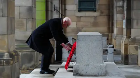 PA Media First Minister John Swinney laying a poppy wreath at the Stone of Remembrance in Edinburgh. He is wearing dark clothes, including a long, dark coat.