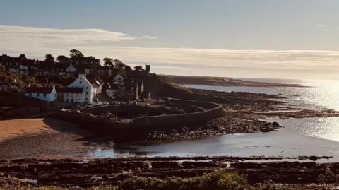 Svend Brown A coastal town. The sun shines on the water with a small sandy beach and rocks by the coast. The little town can be seen in the distance.