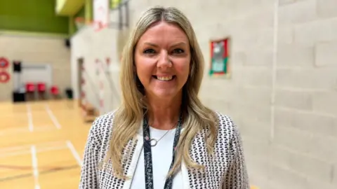 Natalie Johnstone has long wavy blonder hair. She is smiling, wearing a white top with a suit jacket. She has a black Victoria Road School lanyard around her neck and is standing in a sports hall.