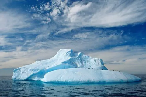 Doug Allan Large iceberg floating in calm ocean water, with a group of penguins standing along the ridge beneath a partly cloudy blue sky.