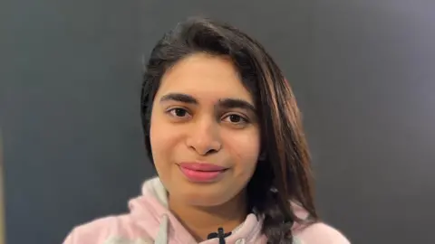 Emma Baugh/BBC Young woman with long dark hair wearing a pink hoody with a smiling face