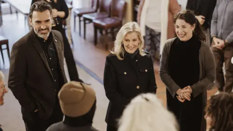 A blonde woman (the Duchess of Edinburgh) in the centre in between a man and a woman talking to other people inside a building.