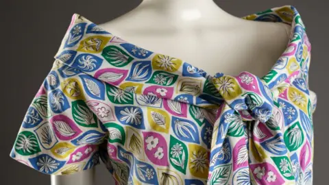 Close-up of the shoulders of a cream mannequin against a dark grey background, wearing a cotton bardot-necked dress with a big tie at the front. The pattern has leaf shapes all over in blue, green, yellow and pink, and within the leaves are other flowers and leaves in white