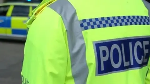 A generic photograph of a police officer wearing a high-visibility yellow jacket with the words police written in capital letters on the back. A police car is in the distance.