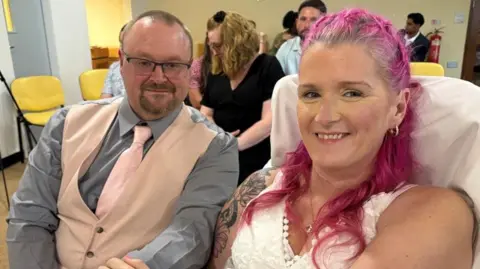 Newlyweds Carla and Craig at a hospital chapel. Carla has bright pink hair and wearing a white dress, Craig is wearing a light pink waistcoat and tie over a grey shirt.