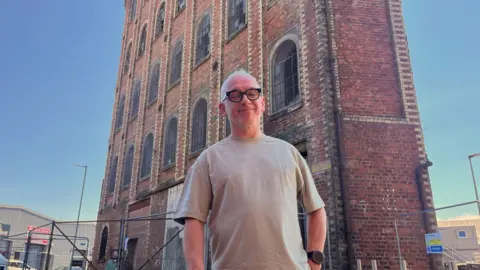 Derek Mitchell stands in front of an old brick industrial building with tall arched windows and metal fencing.