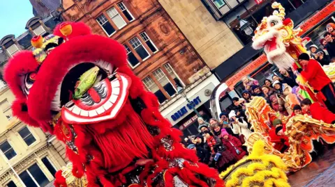 Janina Dolny People dressed in large lion (coloured yellow and red) and dragon (coloured bright red) costumes walking through the Mound in Edinburgh, watched by a crowd of people.