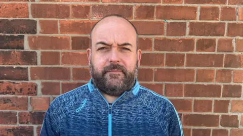 A man with a dark beard is looking directly at the camera. He is wearing a blue sports jacket and is standing in front of a red brick wall
