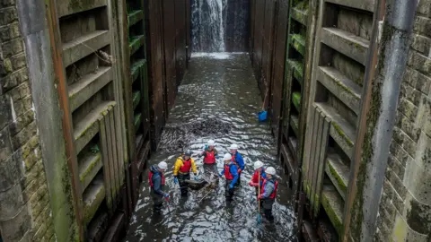 PA Media A group of seven volunteers stand around a shopping trolley which is partially submerged in the water. The trolley contains large waste items.