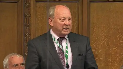 UK Parliament/PA Jim Allister speaking in the House of Commons wearing a suit and tie and green lanyard