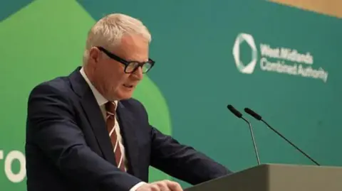 A man in a navy suit jacket with his hands on a lectern and his head down as he reads from notes, mid speech. He is wearing a brown and white striped tie, has white hair and black-framed glasses. A green West Midlands Combined Authority backdrop is behind him.