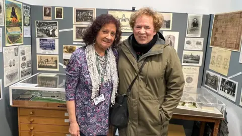 Aisha Iqbal/BBC Two women, older in age, stand in an office space. Behind them are display boards covered in archive photos. They are both smiling.