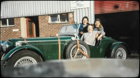 Three women sit in, or perch on, an old-fashioned green car outside a garage. They are all smiling at the camera. The fashion, haircuts and quality of the photograph indicate it is the 1980s.