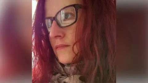 Kelly Anderson is pictured in selfie-style image. She looks away from the camera. She has short red hair, brown glasses and a scarf around her neck.