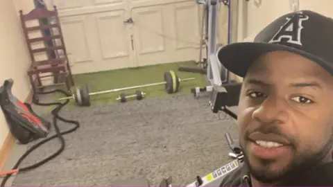 Aaron A selfie of Aaron inside the gym in his garage, with weights on the floor and a rowing machine