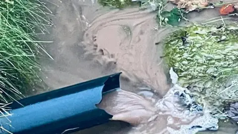 A black pipe depositing brown water into an already flooded area of land. There are some brown leaves near the top of the image.