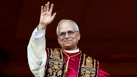 Reuters Pope Leo XIV standing on the balcony of St Peter's Basilica at the Vatican. He is waving and smiling.