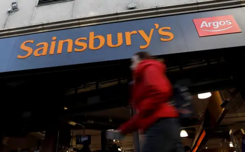Tolga Akmen / AFP via Getty Images The sign to Sainsbury's is seen next to an Argos logo as a person walks past in a red coat, in London in 2019.
