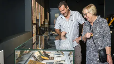 Barnsley Council A room with dark walls which display information boards. A ma with dark hair and a short greying beard wears a short sleeved white shirt with dark stripes. He stands next to a woman with short, blonde hair and glasses who is wearing grey patterned short sleeved blouse and red leggings. Both are looking down at an exhibition case containing old photos and documents.