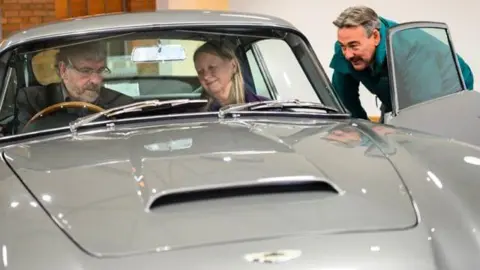 Dominic Fraser/Aston Martin Works John and Susan sit in the car looking at the dashboard as an Aston Martin employee smiles at them from outside the car