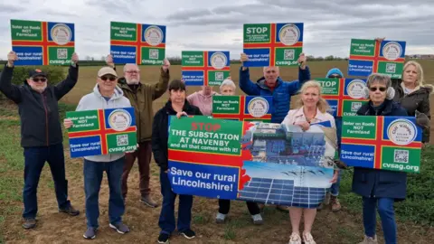Anti-substation protesters stand in a field holding up signs incorporating the green, red and blue Lincolnshire flag with the slogan Save our Rural Lincolnshire
