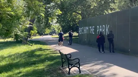 Reclaim the park graffitied on a large green board within Brockwell Park, which is blocking part of the park along a path. To the left of the board is greenery and trees and people walking along the path.