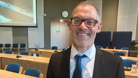 Adam Zerny with short grey hair and beard, with dark-framed glasses, wearing a dark grey jacket, white shirt and blue tie in a council chamber with blue seats behind long wooden tables.