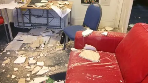 Parts of a ceiling, dropped on a floor, showing a red sofa, large parts of plaster, a blue chair, a desk and paperwork. 