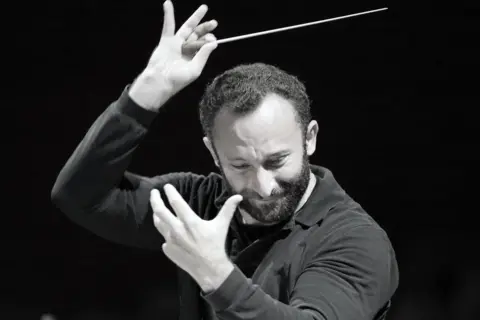 Edinburgh International Festival/Wilfried Ho A black and white photo of a man conducting an orchestra. He is holding a thin baton in his right hand, raised high, while the left hand is positioned slightly lower, with fingers spread in a gesture typical of directing music. He has short, dark hair and a beard and is wearing a dark, long-sleeved shirt. There is a black background.