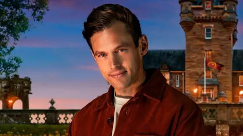 A promo picture of Sam Little smiling in front of the Traitors castle which is a large stone building with a turret. Little has short brown hair which is combed over at the front. He is wearing a red shirt over a white t-shirt. The sky behind him is dark blue with pink wispy clouds.