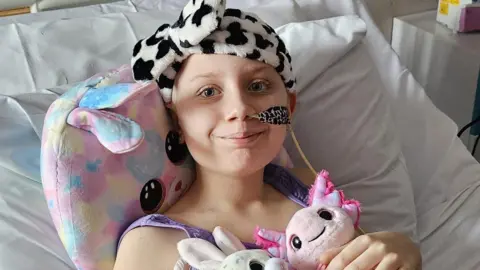 Family handout Megan. She is lying in a hospital bed with three cuddly toys. She has a tube in her nose and a black and white fluffy headband on. She is smiling at the camera. 