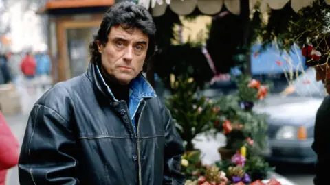 A middle aged Ian McShane in character as Lovejoy, stands with a black leather jacket, blue denim jacked and a black, wavy mullet. He is looking directly at the camera with a serious expression on his face at an outdoor Christmas market, with wreaths and decorations on the stall he is standing in front of. In the background, the bonnet of a grey, second generation Vauxhall Astra can just be made out, despite being out of focus.