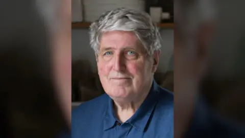 Tom Maine John Maine looks directly at the camera as he poses for a headshot. He has short grey hair and blue eyes. He is wearing a french blue linen collared shirt and the background behind him is blurred.
