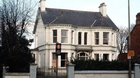 A white building with a black roof. It has lots of windows in the front and a dark gate with grey pillars at the entrance. There is a pedestrian crossing outside the gate.