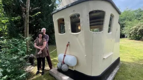 Wide-angle lens photograph of Richard and Abi stood next to their cabin - a wooden and steel structure painted cream with two small windows on each side. On the front of the cabin are buoys and fenders, and it's 'tied up' to a black mooring post. It's in a garden, with green trees and bushes along the edges of green grass. Richard and Abi are stood with their arms around each other. 