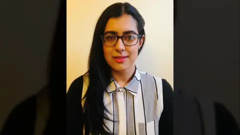 Yasmin Shabir A woman with long dark hair is shown in a simple portrait against a plain light‑coloured wall. She is wearing glasses and a black‑and‑white striped collared shirt.