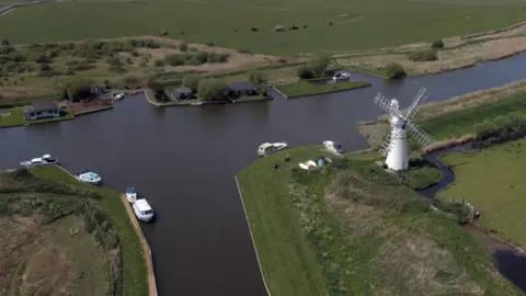 Martin Barber/BBC The Broads runs between three fields. White and blue boats are stationary on the edges of the stretch of water. On the right, a white windmill sits on a section of land. 