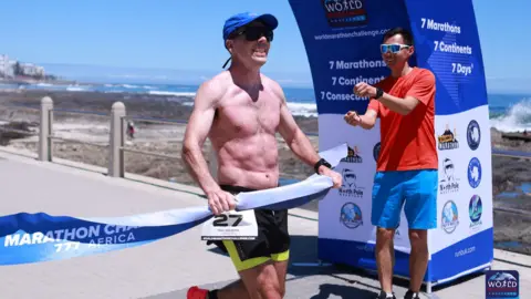 World Marathon Challenge/Runbuk Paul crossing the finish line in Cape Town. He's shirtless with a blue cap on. He's got a hold of paper line indicating he's finished first. He's smiling ahead of him. The sea can be seen in the background.