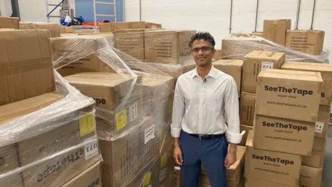 Dr Rajan Bhojwani standing in front of stacks of boxes in a warehouse. The boxes display the brand name See The Tape.