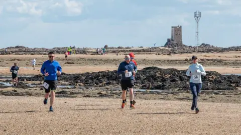 Philip's Footprints Several runners crossing a beach with rocks and a castle in the background.