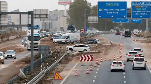 Kai Försterling/EPA-EFE/REX/Shutterstock The A3, the main road between Valencia and Madrid, is partially closed, with some vehicles stuck in mud and central reservations visible. Some cars can pass a cone-shaped section of road in both directions. There are motorway signs on the road indicating Valencia Airport and Barcelona