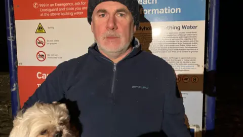 A man standing in front of a sign with various information about a beach. He is wearing a navy fleece and hat and holding a small white dog.