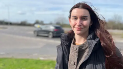 Alison Holmes looking into camera with the junction in the background. Alison has long brunette hair and is wearing a dark jacked and light brown jumper.